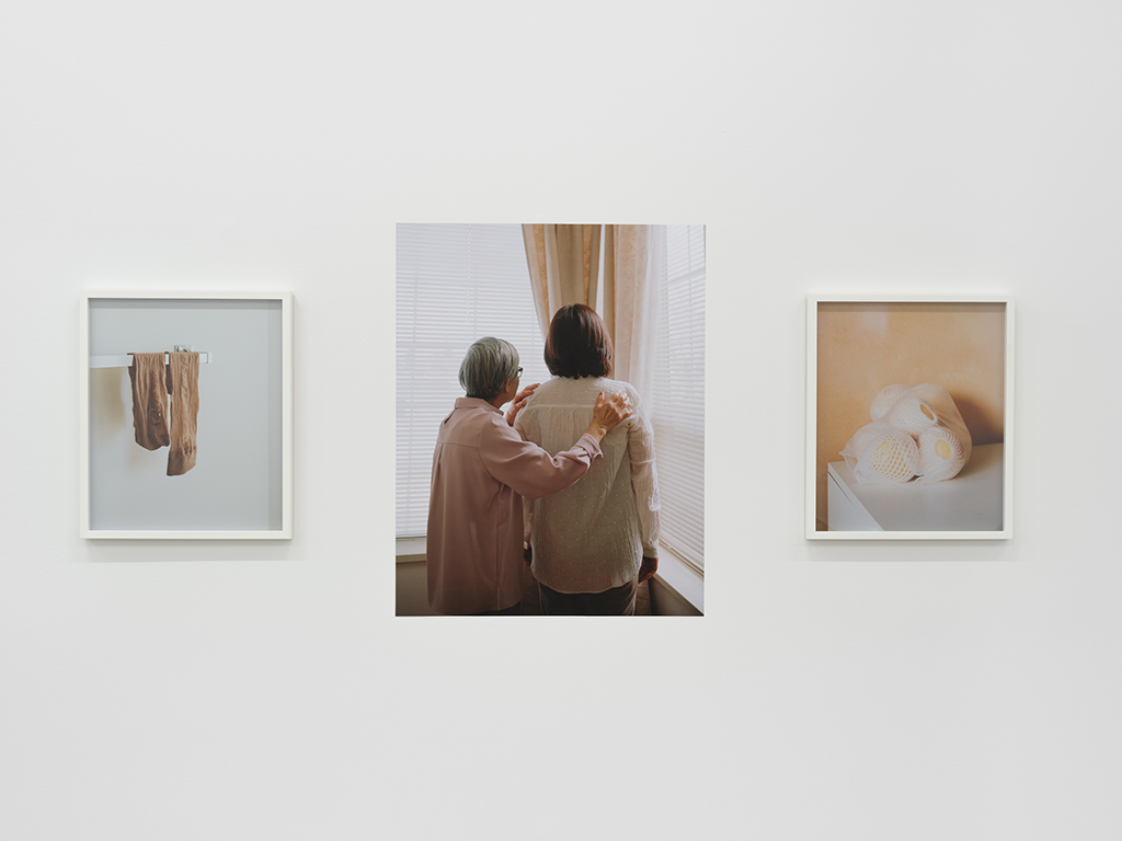 An installation view of Gloria Wong’s <em>Socks, Ngan and Pears</em>, three complementary images that depict, from left to right, a pair of brown nylon socks, the artist’s grandmother and apple pears wrapped in white foam netting.