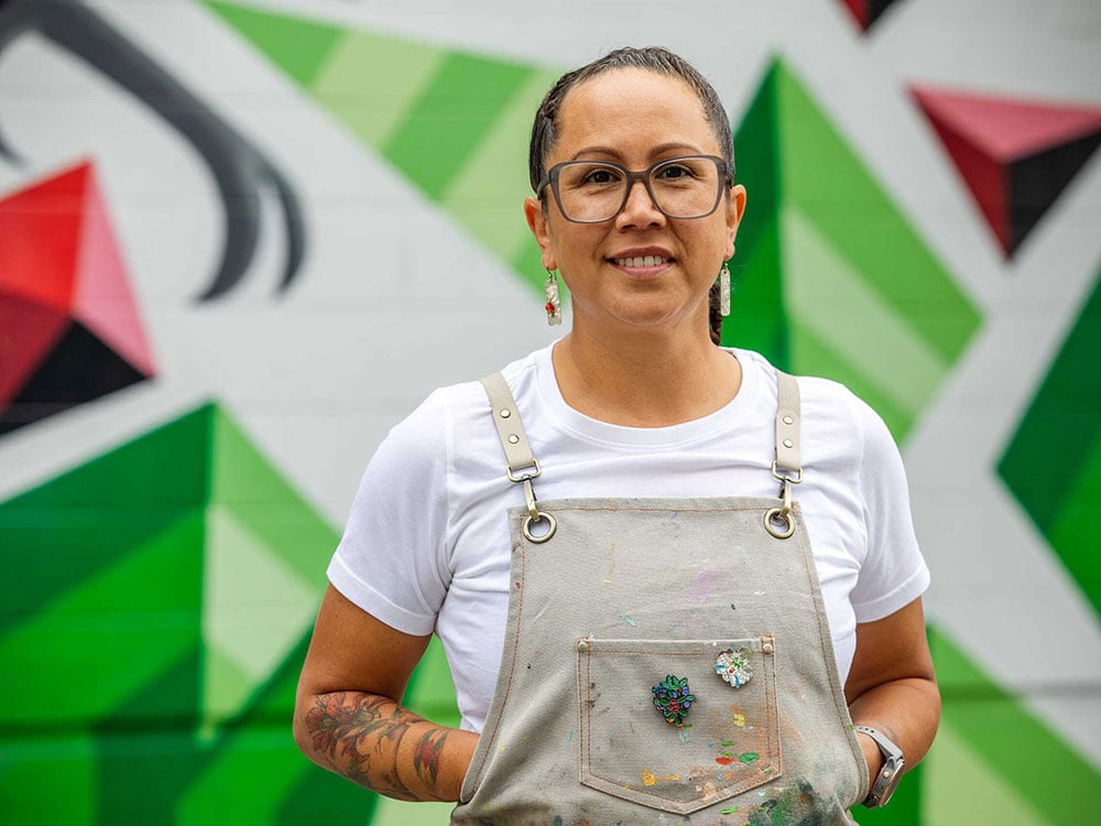 Xémontalót Carrielynn Victor is wearing glasses and overalls. Her hair is braided and she is looking at the camera, smiling. Behind her is a mural with green, white and red colours.