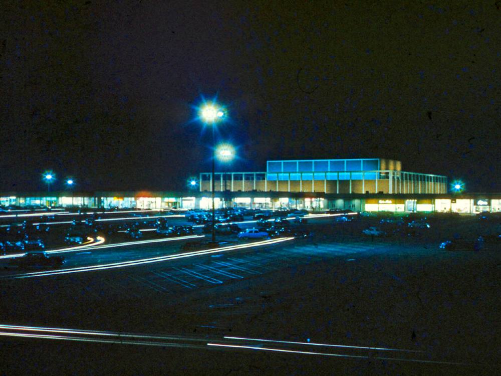It’s night and a boxy department store rises behind a strip of connected shops, all aglow in the dark. In the foreground is a large parking lot with a smattering of cars. The scene is lit with a few tall lamps.