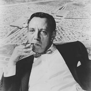 A black and white portrait of a man leaning back in his chair at an angle. Holding a pen in his mouth, he appears to be in deep thought. He is in a suit with a bowtie. There is a poster behind him of an aerial view of a shopping mall.