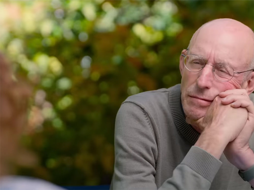 Michael Pollan is seated at a table, resting his cheek against clasped hands while interviewing a person across from him. The back of their head is partially visible. Pollan is seated against a lush green background. He is wearing glasses and a grey shirt.
