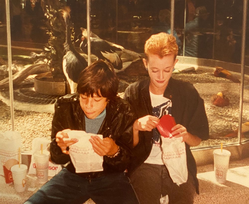 Two teens are looking down at the snacks in their hands. They are Dorothy Woodend (right) and her friend Rueben (left). They are sitting on a ledge in front of an atrium at West Edmonton Mall in the 1980s.