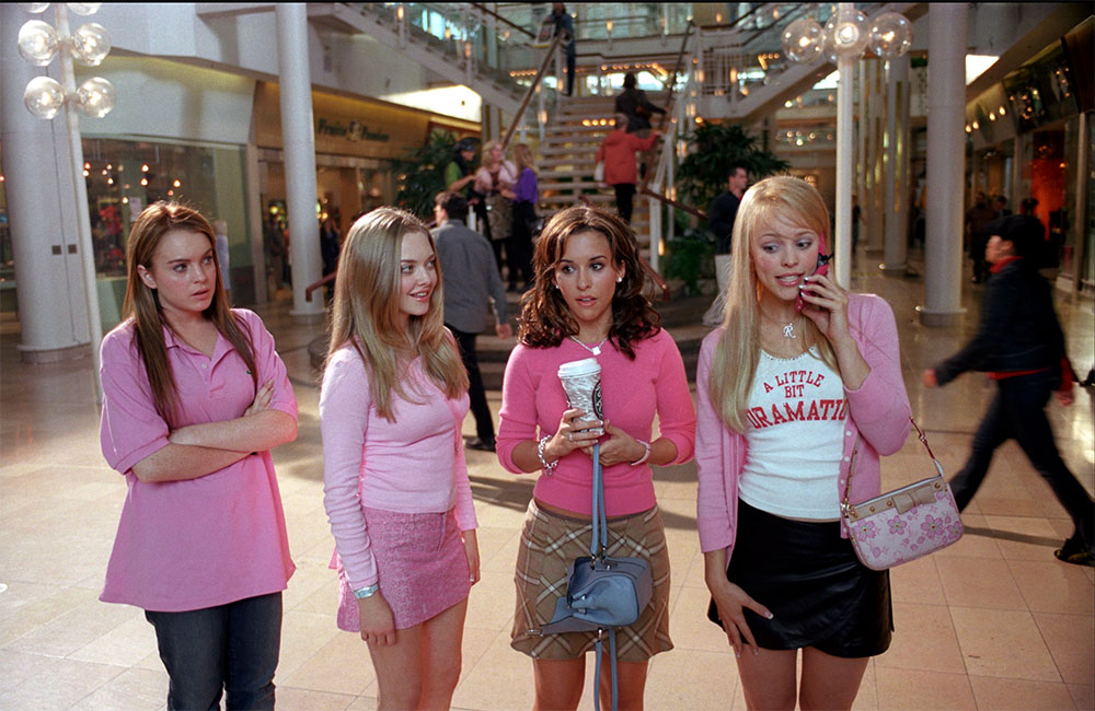 Four teen actors are standing next to each other, all of them wearing pink with various expressions on their faces. Behind them is a hallway and staircase in Toronto’s Sherway Gardens.