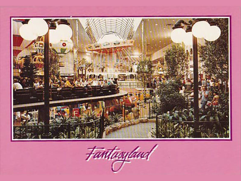 A vintage postcard features a pink border with the word “Fantasyland” in stylized script below a colour photograph of an atrium at West Edmonton Mall. The multi-level atrium is busy with diners, shoppers and amusement park rides.