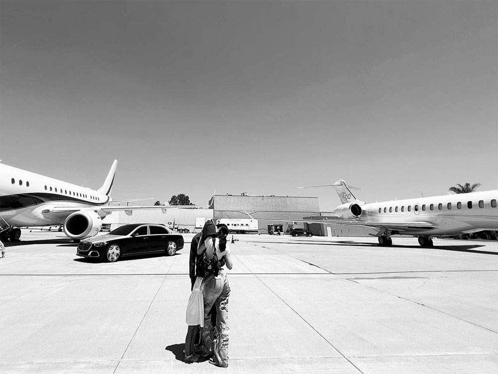 A black and white photo of Kylie Jenner embracing her partner Travis Scott on the tarmac of an air terminal. They are standing between two private jets.