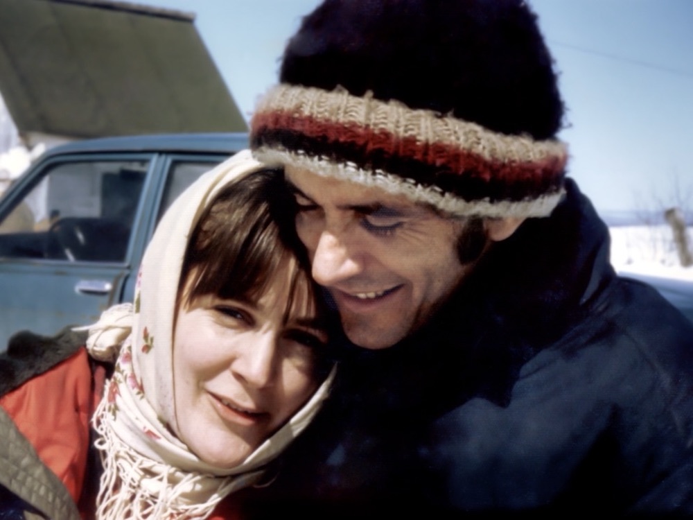 A colour photo of Audrey Schirmer and Martin Duckworth depicts the young couple on a sunny day in the winter. Schirmer is wearing a scarf over her head and a red jacket. Duckworth is in a toque and navy winter coat.