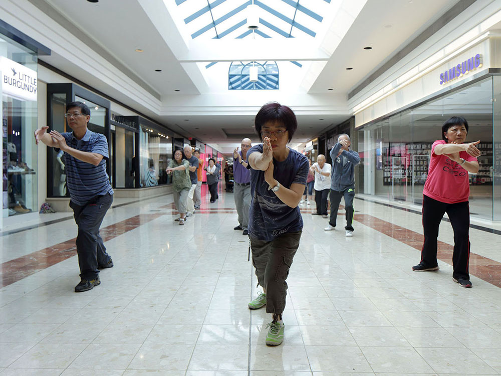 A group of Chinese seniors in the hallway of a shopping mall doing tai chi. On the left and right are storefronts like Little Burgundy and Samsung, which have not yet opened at this morning hour. 