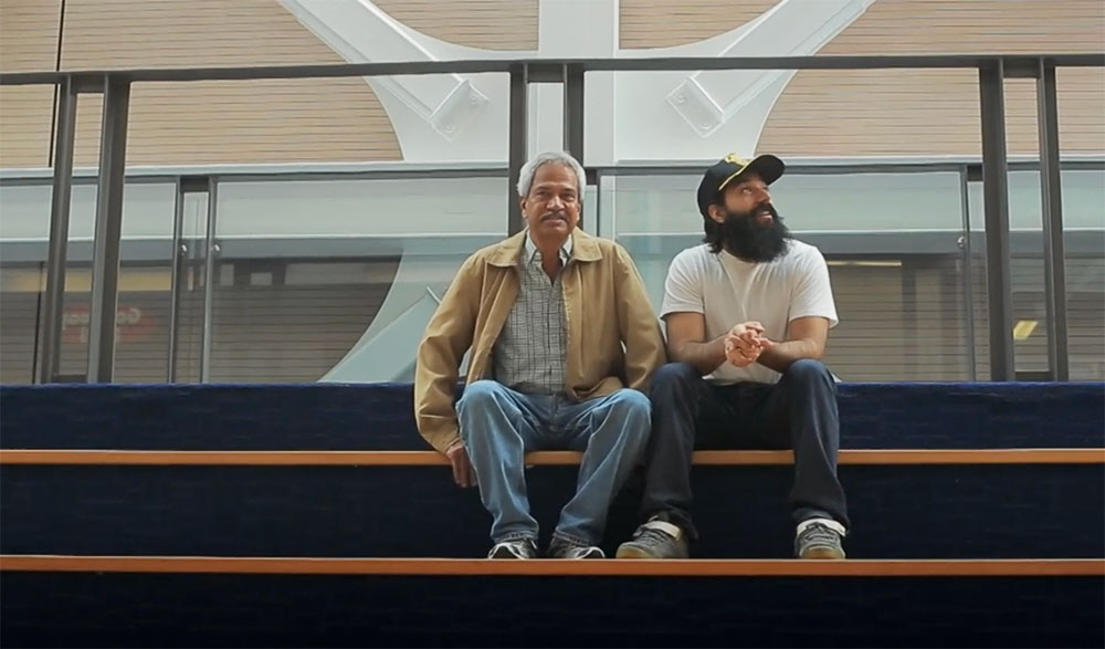 Inderjit Mahal and Manny Mahal sit in in the empty bleachers of a local hockey rink. The seats are navy blue. Inderjit and Manny are seated near the top back rows. 