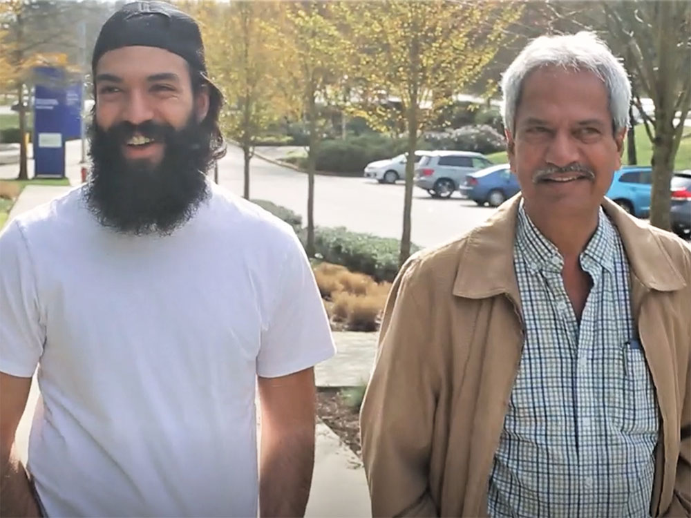 Manny Mahal is wearing a white t-shirt and backwards black ball cap. He has a long black beard and is looking into the distance, smiling. His father, Inderjit, is to his left. He’s wearing a tan jacket over a plaid shirt. He has short grey hair.