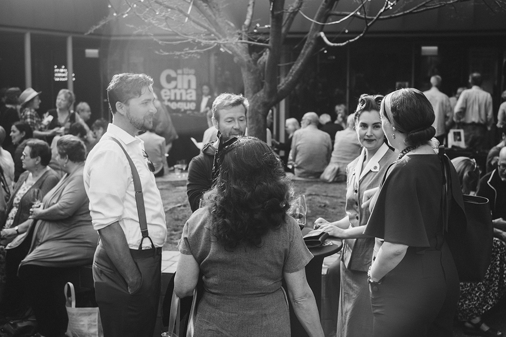 A black and white photo of The Cinematheque’s lobby, where people are gathering and chatting. In the foreground, a cluster of younger adults stands around a table on which there are glasses of red wine.