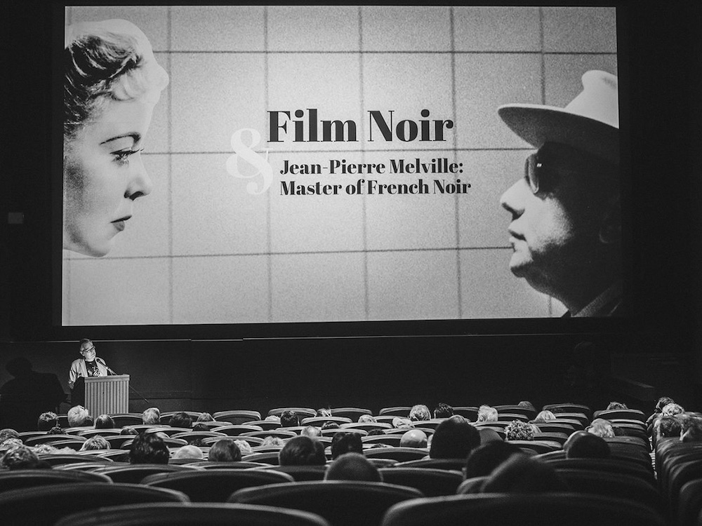A black-and-white photo of a screening room at The Cinematheque is shot from the back of the theatre. One can see rows of audience seating and a person speaking at a podium onstage to the left of the screen. The silver screen reads “Film Noir and Jean-Pierre Melville: Master of French Noir.”