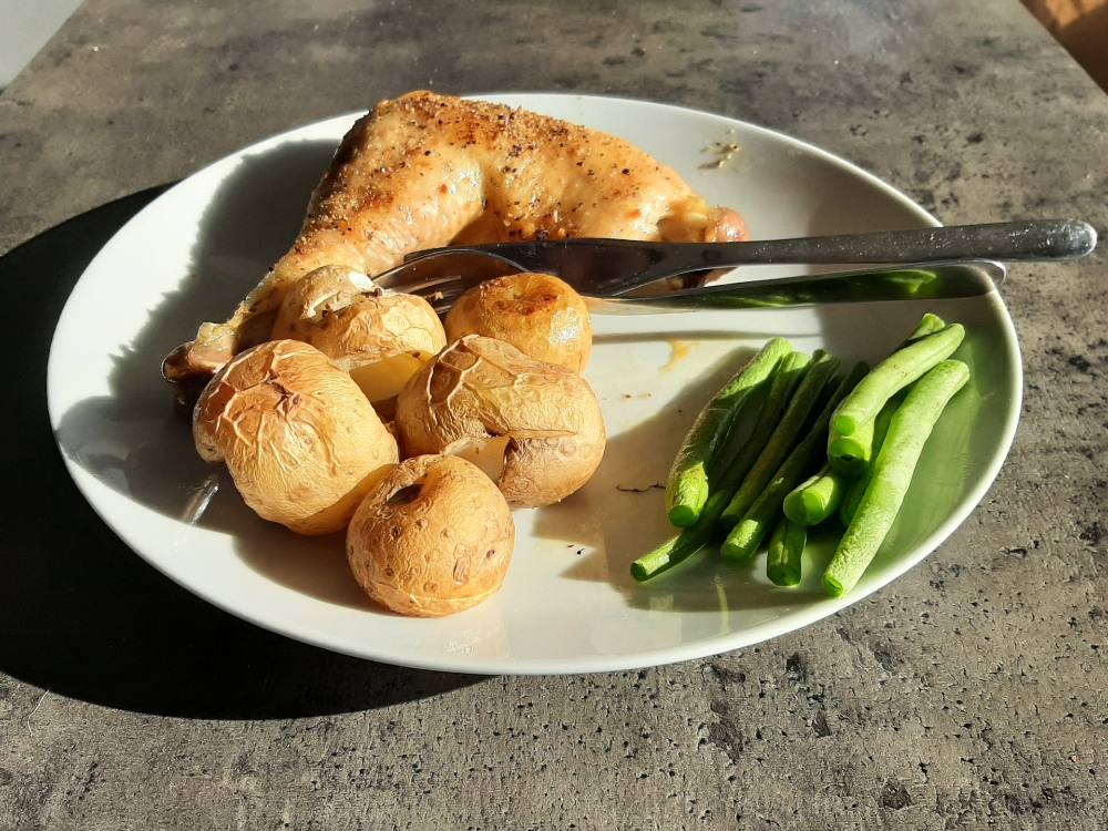 A sunny plated dinner features a baked chicken leg, baked potatoes and friend green beans on a white plate.