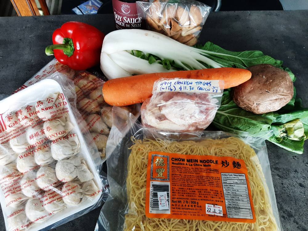 A selection of groceries sits on a grey countertop. A red bell pepper, orange carrot and bright bunch of bok choy cross the top of the frame. They are nestled between a bag of fortune cookies, a package of chow mein noodles, frozen packages of dim sum, and chicken thighs.