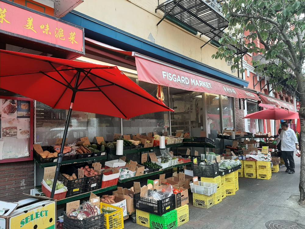 A sidewalk view of Victoria’s Fisgard Market shows cases of produce stacked on outdoor shelves under bright red awnings.