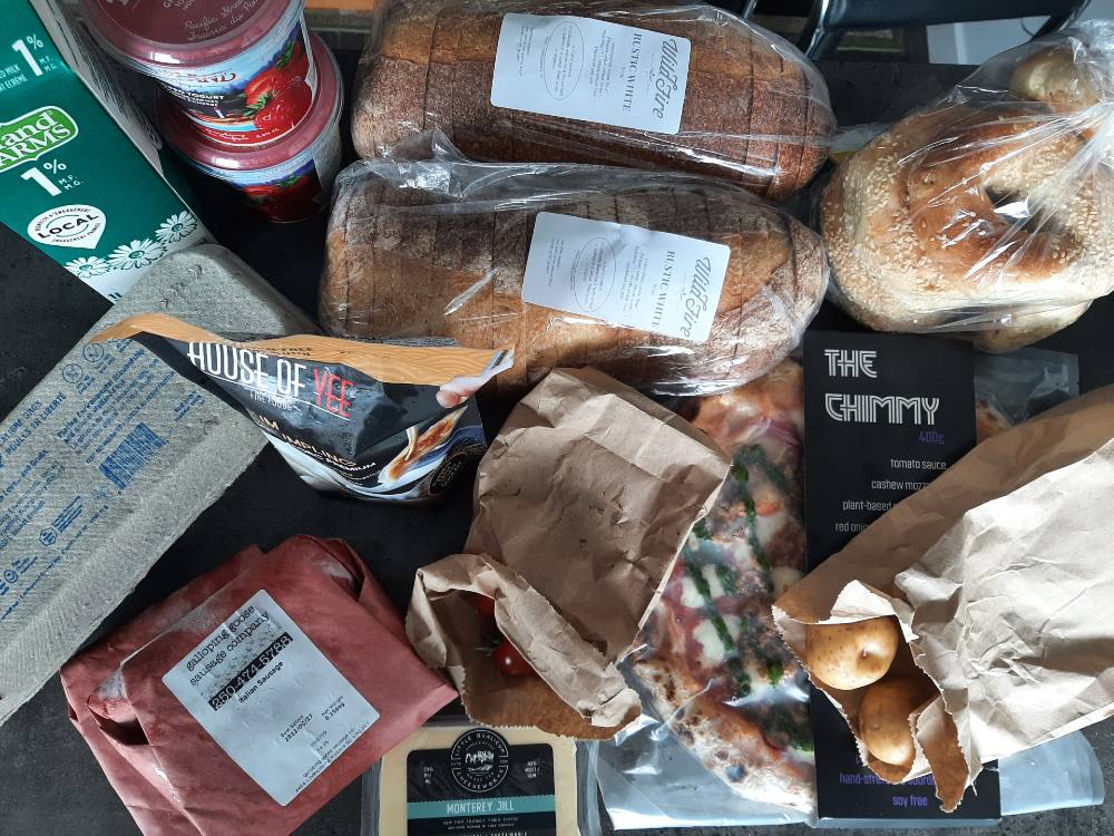 A bird’s-eye view of a selection of groceries from For Good Measure in Victoria features two loaves of brown bread, a green carton of Island Farms milk, strawberry yogurt, a carton of eggs, and several packaged items to the bottom right corner of the frame.