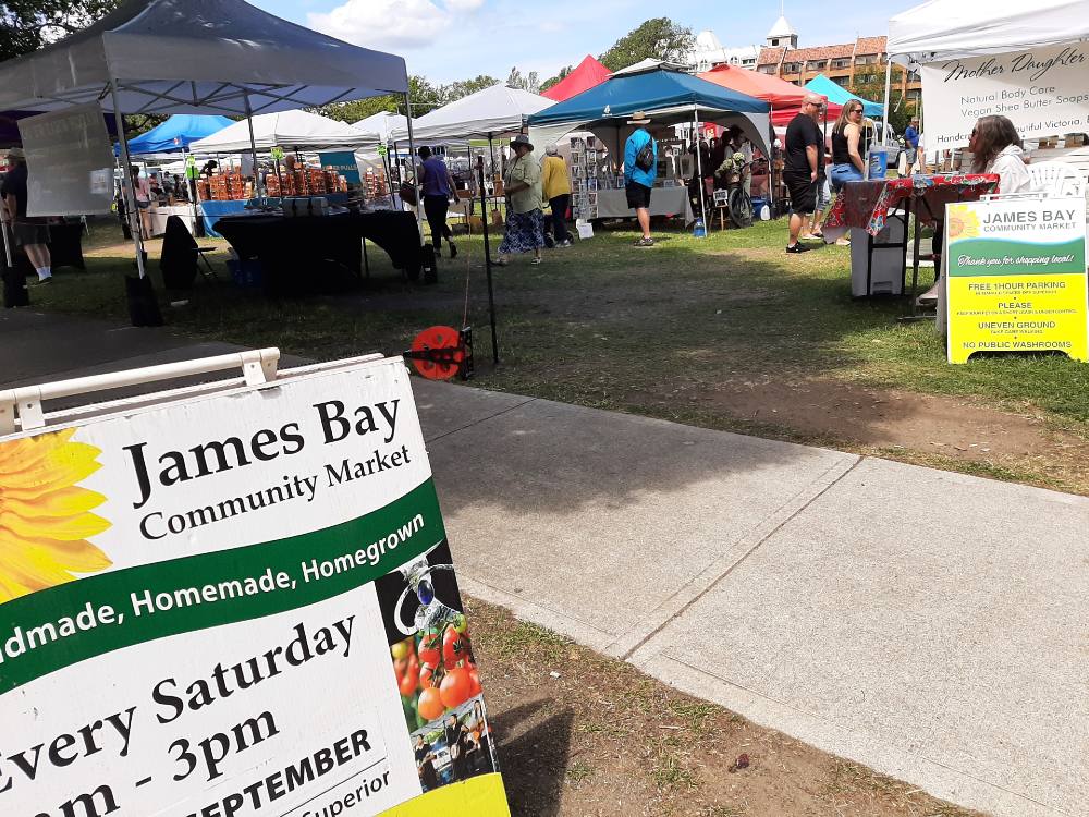 The entrance to James Bay Market, a farmers market in Victoria, features a colourful sandwich board that reads "James Bay Community Market.” There are several tented vending stalls in the background.