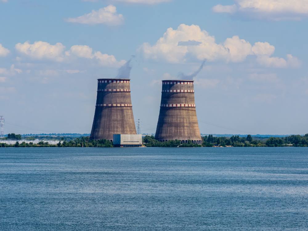 Two cooling towers of Zaporizhzhia Nuclear Power Station near the city of Energodar, southeastern Ukraine. The water in the foreground and sky in the background are blue. The towers are two brown towers next to each other with three red and white stripes.
