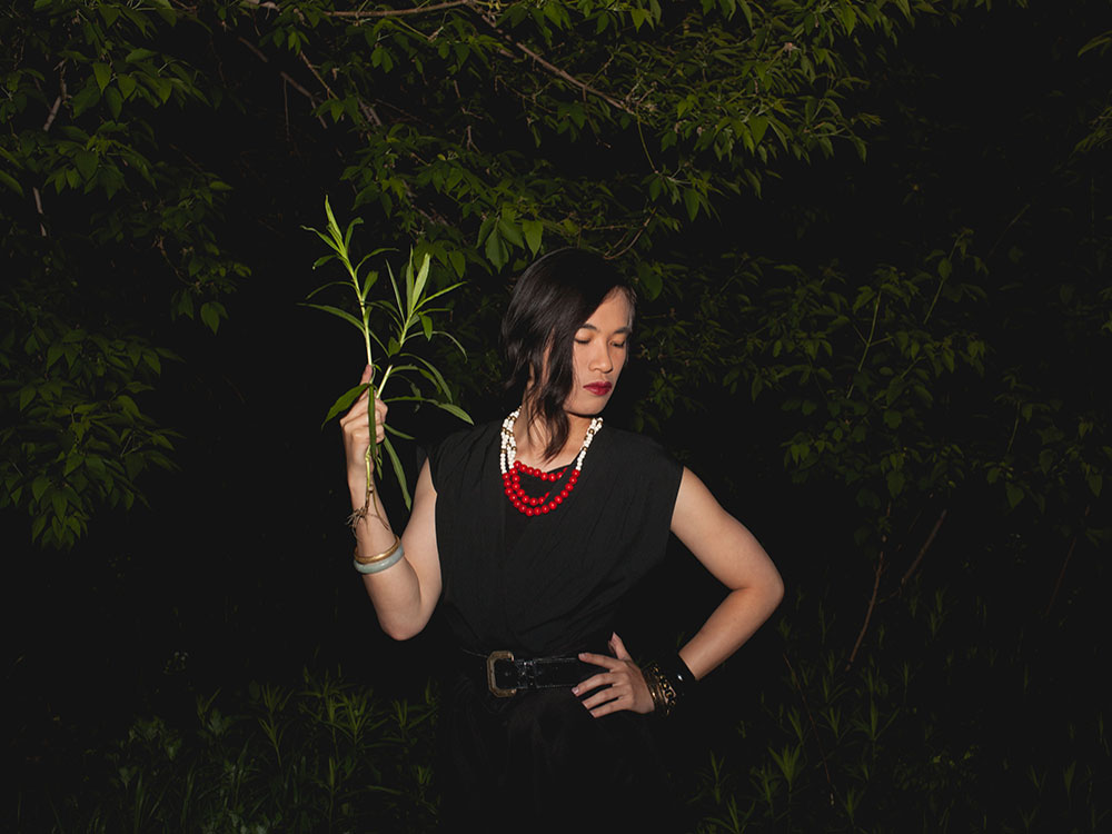 Kai Cheng Thom is standing against a backdrop of leafy green trees at night. She is holding one branch in her left hand. Her right hand is on her hip. She is wearing a black dress and a necklace with red and white beads. She is looking down towards the right side of the frame.