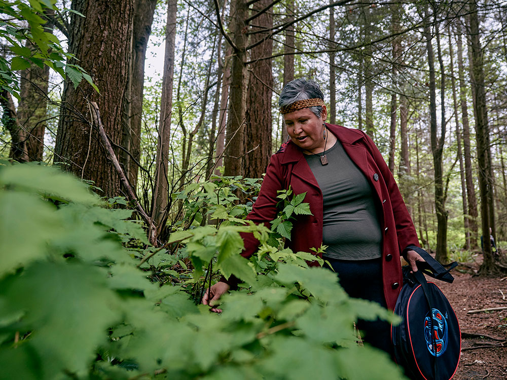 A person wearing a red jacket and a headband stands in a forest, picking a berry from a bush.