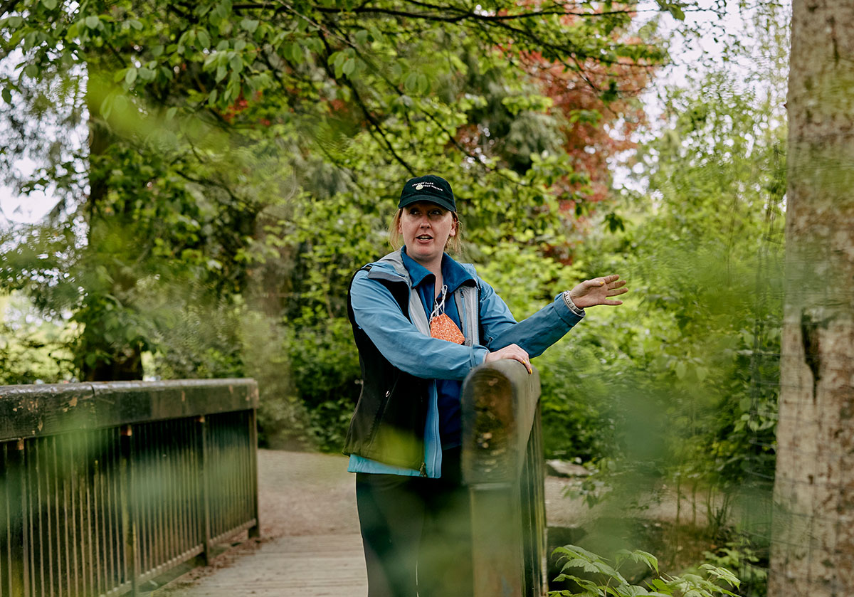 A person stands on a bridge in a park, wearing long sleeves and a vest, talking and pointing with their left hand. 