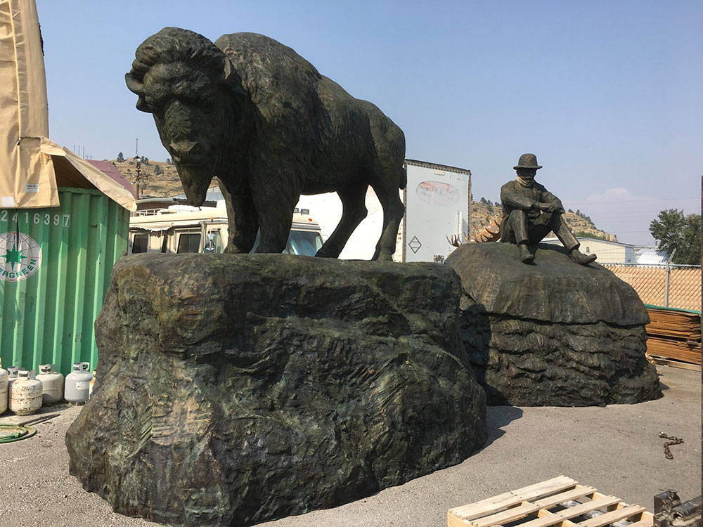 Two bronze sculptures, one of a bison and the other of man sitting on a pile of furs wearing a western style hat.
