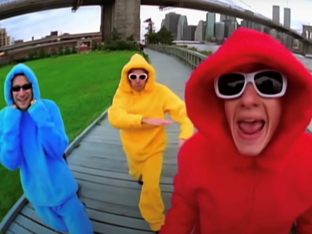 Three people in fleece suits amble across a boardwalk along the Hudson River in New York City. One can see the Manhattan skyline in the background. From left, Adam Horovitz is in blue, Adam Yauch is in yellow and Michael Diamond is in red. They are all wearing sunglasses.