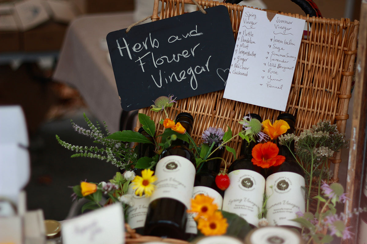 A basket of tall dark glass bottles containing herb and flower vinegar lie beneath a chalkboard sign. The bottles are decorated with colourful wildflowers.