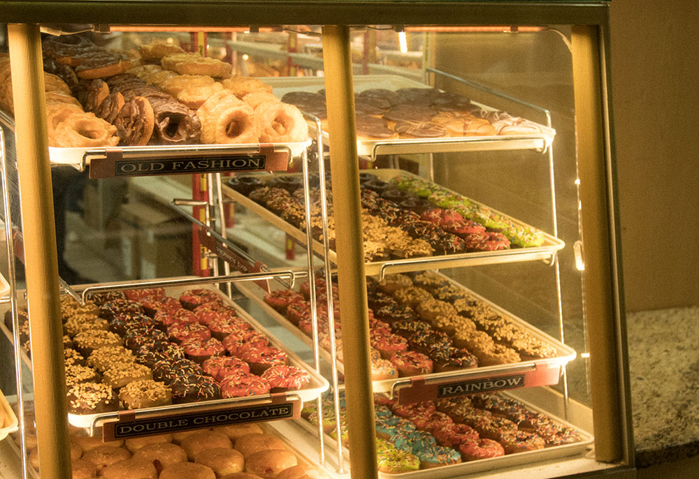 Seven trays of doughnuts are displayed behind a glass cabinet. Flavours include ‘old fashioned,’ ‘rainbow’ and ‘double chocolate’. The glazed sugar and sprinkles glistens in the soft lights. 