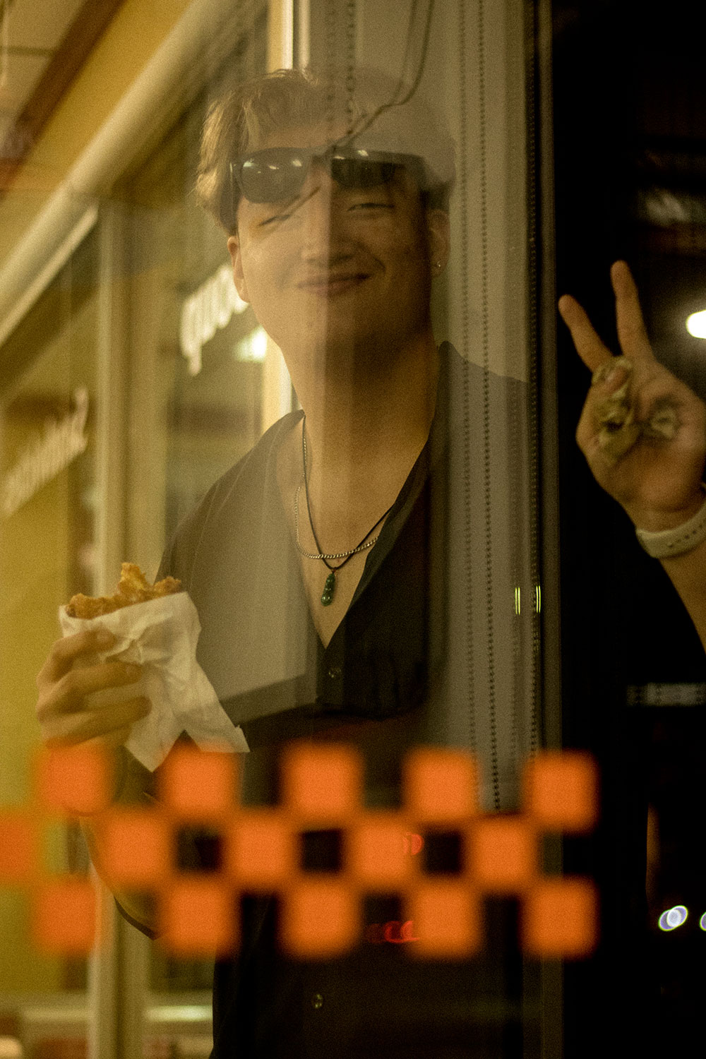 A young man eating a honey crueller donut makes a peace sign towards the camera through the window, his sunglasses precariously balanced on his head and a goofy smile on his face. 