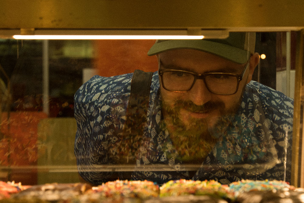 A man wearing glasses and a baseball cap looks through the glass cabinet at a row of rainbow sprinkled doughnuts. 