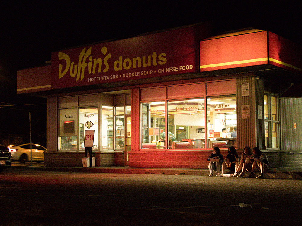 Four teenage girls sit on the curb outside Duffin's Donuts. They are framed by the bright lights of the diner, and lit up by the headlights of a car driving past. 