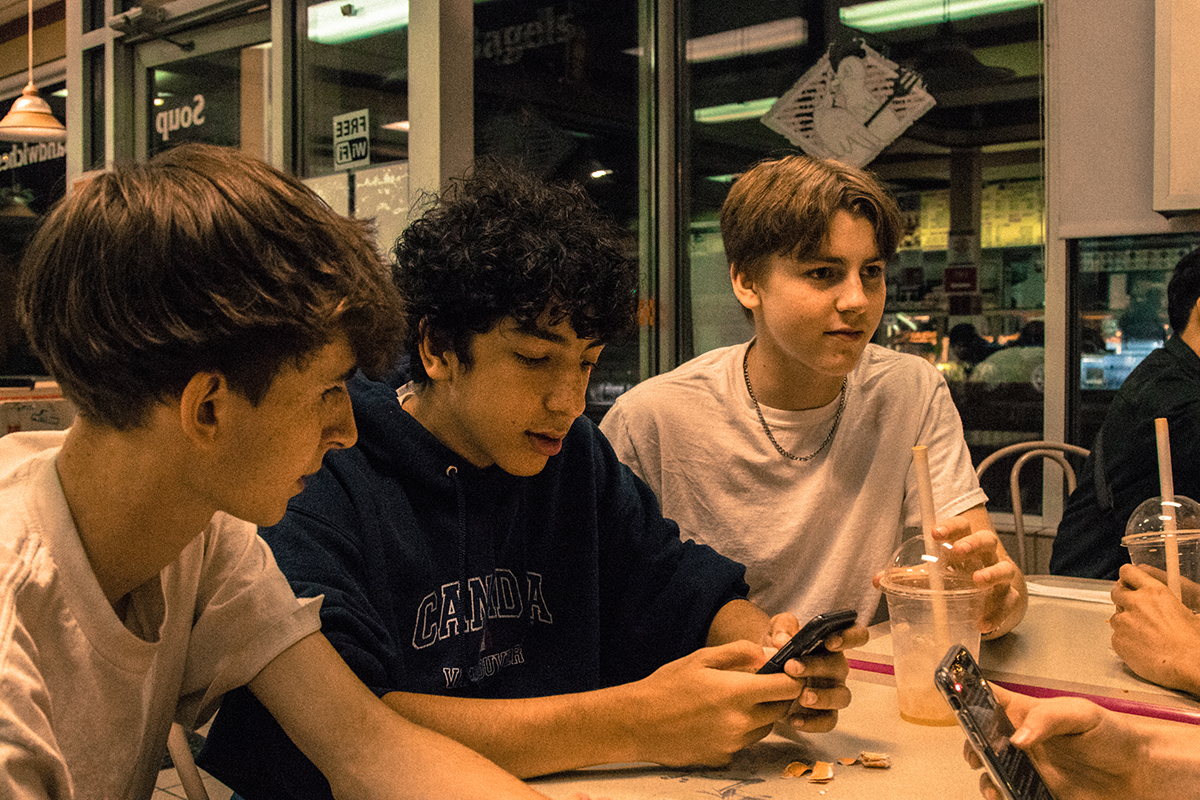 Three teenage boys gather around a table at Duffin’s Diner, two are looking at a phone while the other — sporting a 1990s Leo Dicaprio look — looks to the other side of the table where two other friends (off-camera) are talking. 