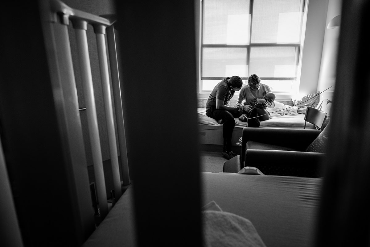 Parents wearing masks sit in a hospital bed, cradling a baby. The photo is taken through a door window and we are looking at this intimate scene from a distance.