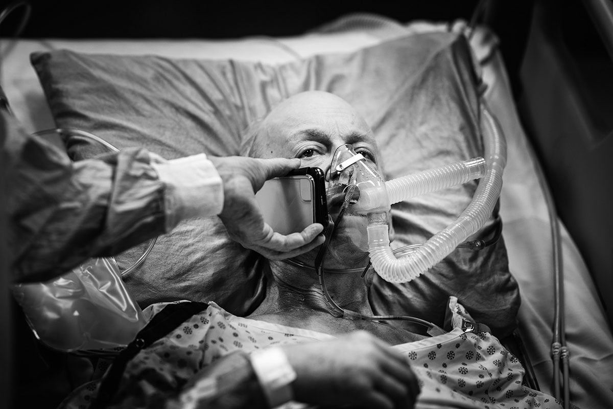 A nurse holds a phone to the ear of an elderly man in a hospital bed. There are thick tubes coming out of his mouth in preparation for his bronchoscopy.