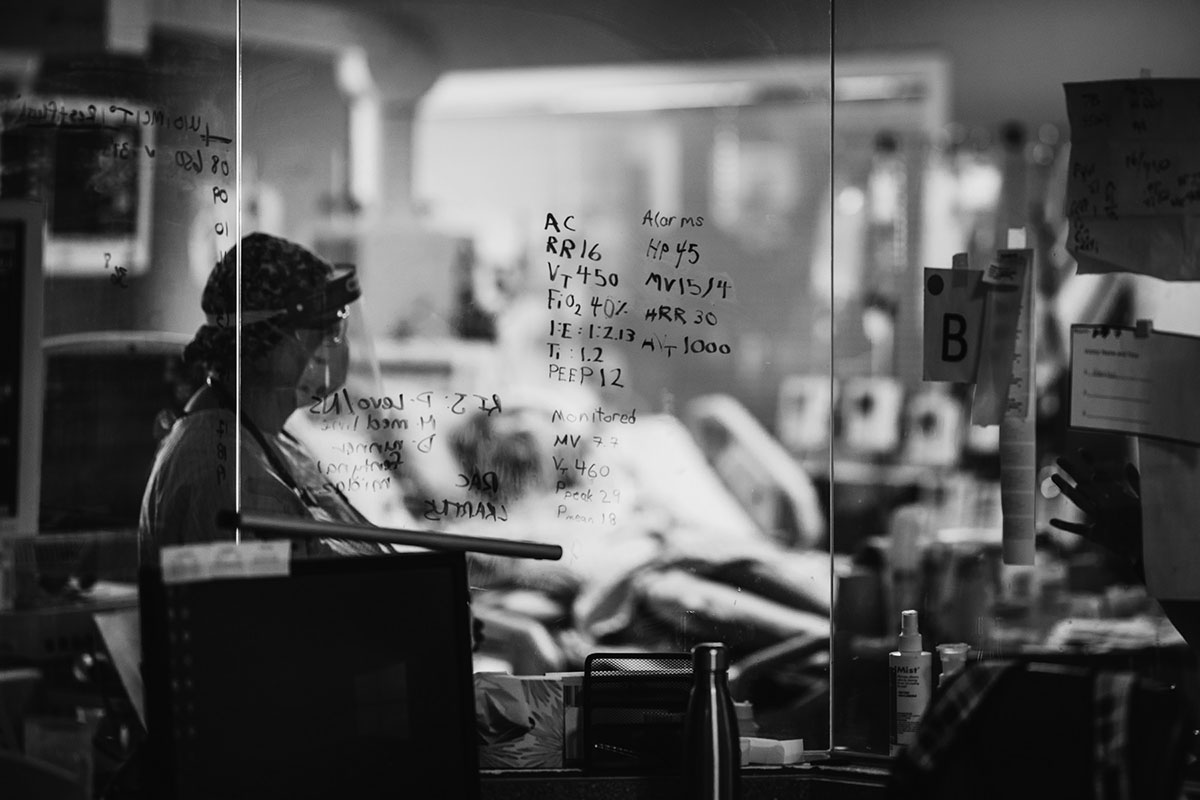 A nurse wearing a mask and face shield in a hospital’s isolation room, seen through a window with ventilator settings written on its surface.