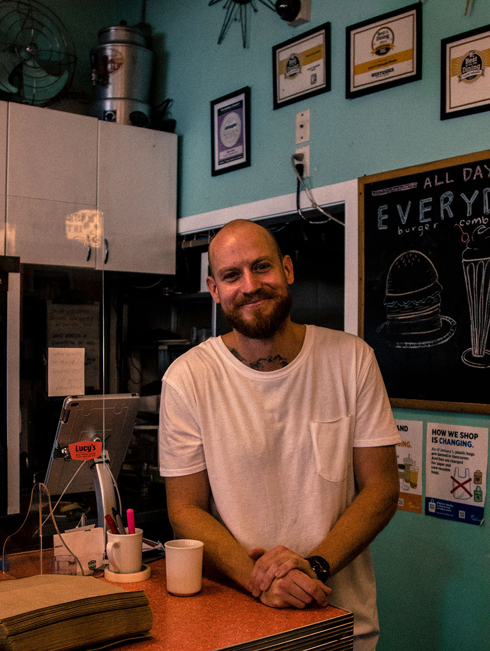 A man wearing a white T-shirt, from the collar of which peeks a neck tattoo, leans against the counter and smiles at the camera. 