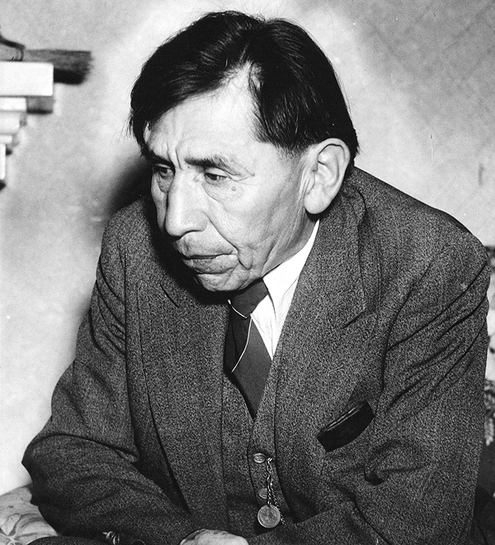 A black and white portrait of August Jack Khatsahalno wearing a suit jacket and tie. He is seated at a table, looking down towards the left of the frame, his expression serious.