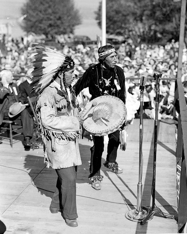 A black and white archival photo depicts August Jack Khatsahlano getting ready to perform a dance at the rededication ceremony for Stanley Park. Khatsahlano is in black and he is standing next to a person holding a drum. The two are standing onstage with dignitaries in suits behind them. A crowd is in soft focus in the background.