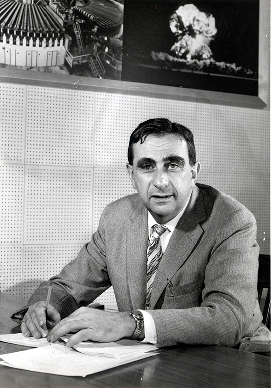 A black-and-white photo of a man in a wool suit. He is sitting down. His hands rest on sheets of paper on a table in front of him. Behind him, there is an image of what appears to be a nuclear explosion tacked to the wall.