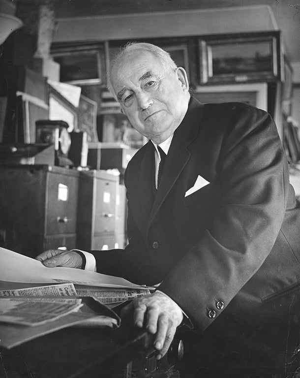 A black and white archival photo of Major James Skitt Matthews. He is wearing a black suit and seated in an office. There are filing cabinets in the background and Matthews is handling paper documents at a desk.
