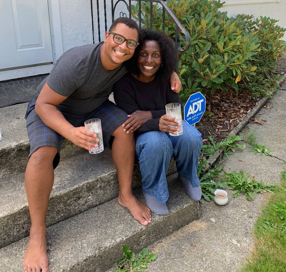 Harrison Mooney is wearing glasses and is holding a glass of water. He is to the left of the frame and seated on the grey concrete steps of a house with his mother, Trinika, right. Trinika is wearing a black top and jeans. They are both looking at the camera, smiling.