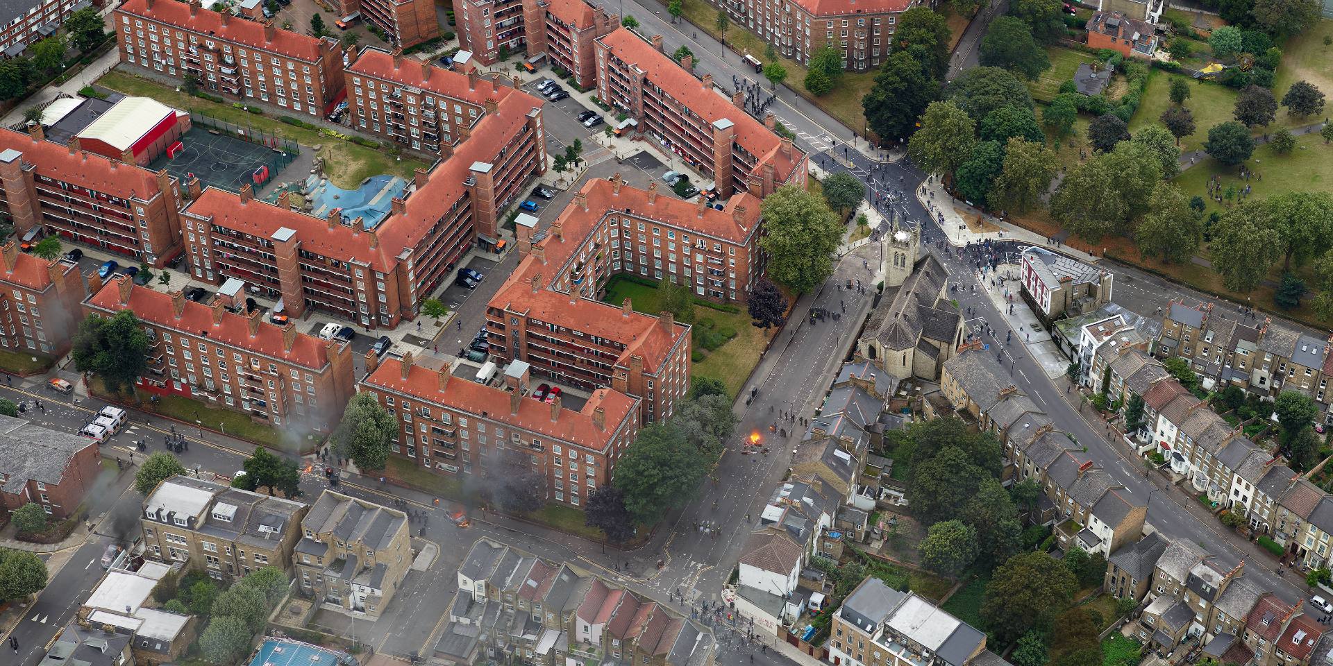 An aerial shot of the Pembury Estate housing complex in red brick and the surrounding streets. Small fires are igniting in the streets, emitting plumes of black smoke. 