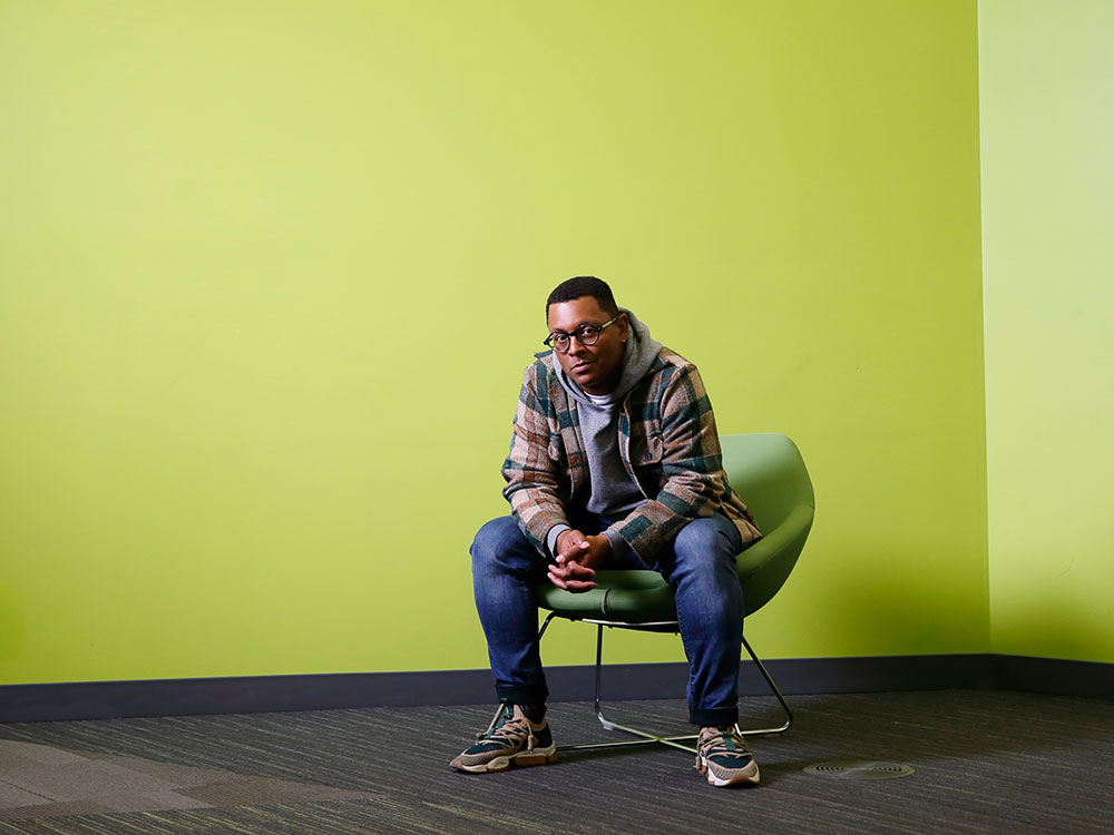 Harrison Mooney is seated on a green chair against a bright green background in the Vancouver Public Library’s Inspiration Lab. He is wearing glasses, a grey hoodie under a plaid jacket, jeans and sneakers. He is looking at the camera with a serious expression.