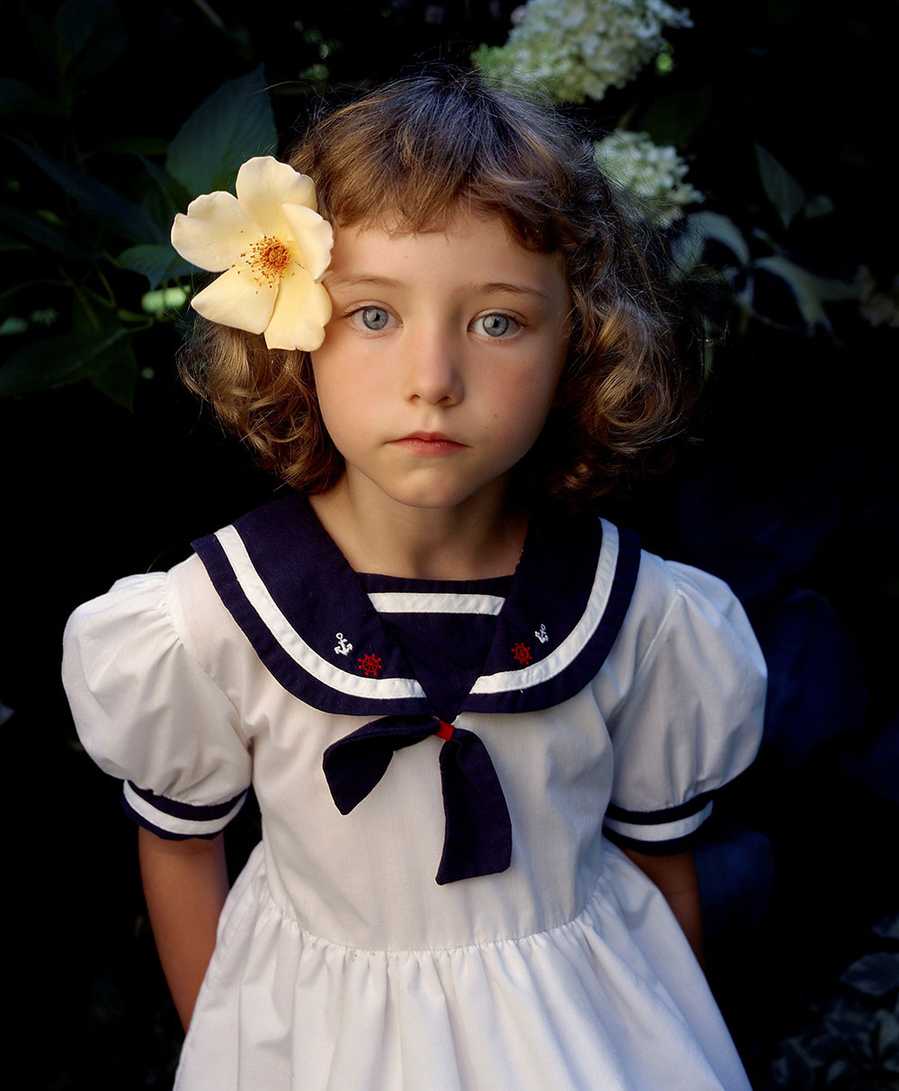 A young girl in a sailor dress looks up to the camera with a blossom tucked behind her ear.