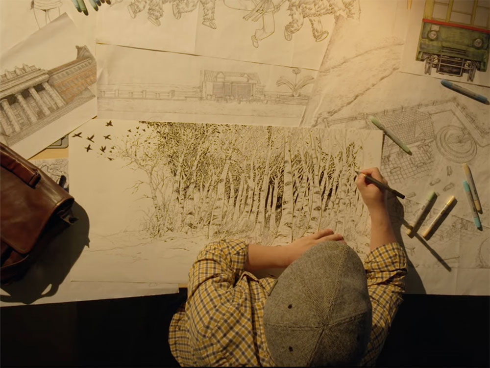 An overhead shot of the artist Daxiong at a drawing table. The background is dark and Daxiong is wearing a grey cap and a beige button-down shirt with a plaid print that matches the beige table on which a collection of his large-scale illustrations is arrayed.  