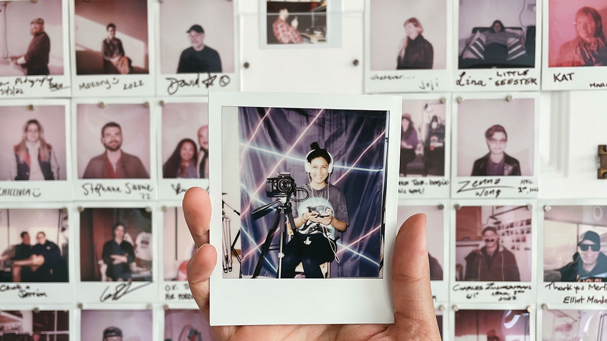 A hand holds a Polaroid of a woman sitting by a video camera, wearing large headphones and smiling. In the background, there are a lot more polaroids.
