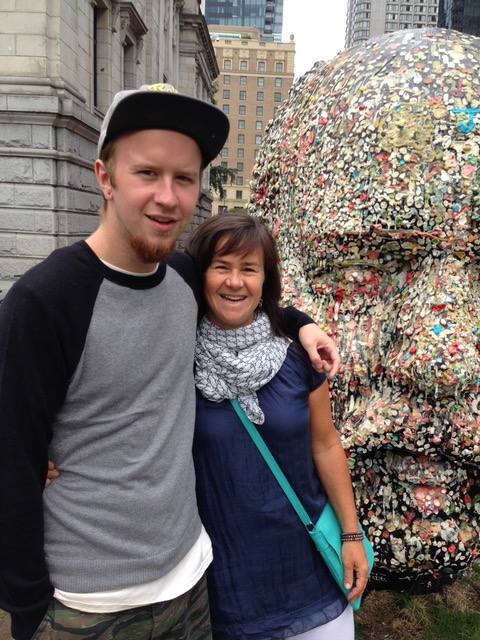 Tara McGuire, right, is wearing a white scarf, dark blue top and turquoise purse. Her son Holden, to the left of the frame, is standing with his arm around her. He is wearing a black and grey henley and ball cap. They are both smiling and standing near a sculpture in the shape of a person’s face outside the Vancouver Art Gallery.