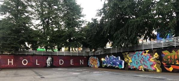 Two stacked photo depict a graffiti artist at work on a piece of artwork with light pink, blue and yellow tones. Below the photo of the artist is a photograph of a wall memorializing Holden. The name “Holden” appears in white text across a burgundy painted wall with a black and white painting of Holden’s face. He is smiling.