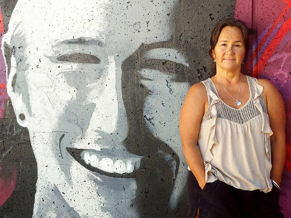 Tara McGuire, right, is wearing an oyster grey sleeveless top with ruffles. Her brown hair is pulled back. She is looking directly at the camera with a warm expression on her face. She is standing against a wall depicting her late son Holden, who died when he was 21. His face is painted in black, white and grey. He is wearing a backwards ball cap, he has a piercing in one ear and he is smiling. 