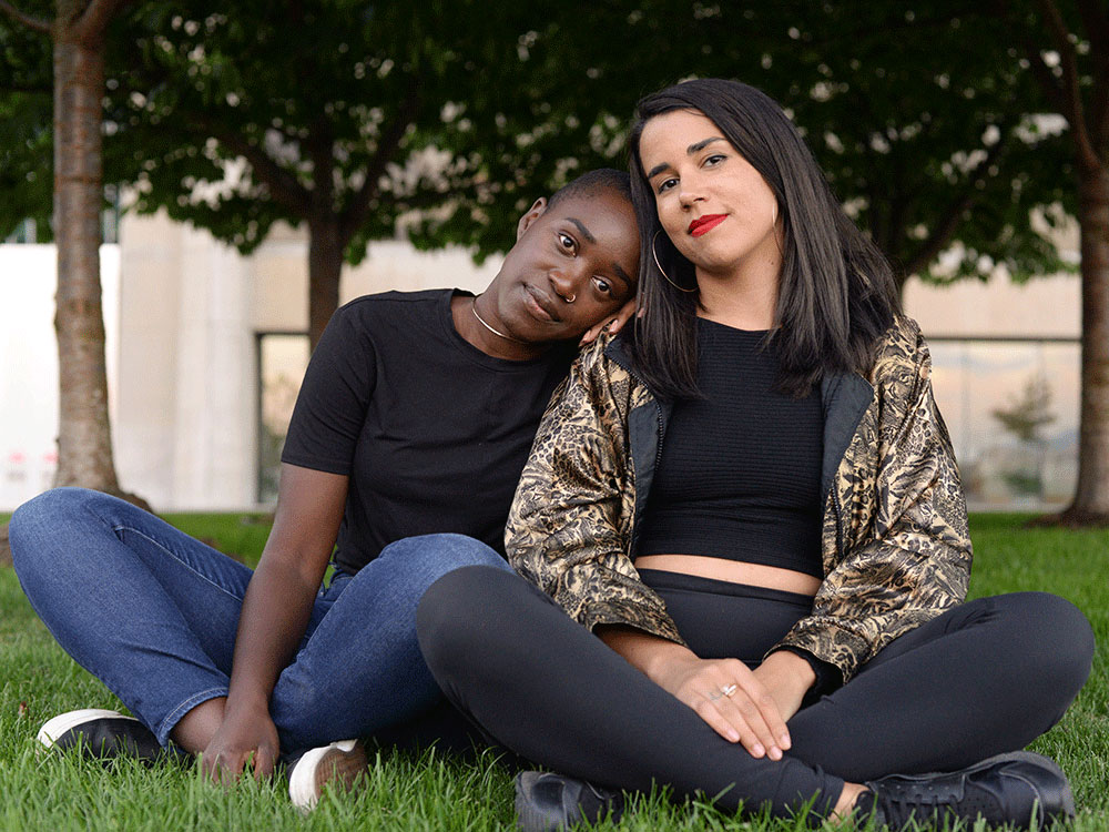 Two people are seated cross-legged on grass. Nic Wayara, left, is wearing a black top and blue jeans. She leans her head on the shoulder of Premala Matthen, right, in a black top and gold jacket. They are both looking serenely at the camera.